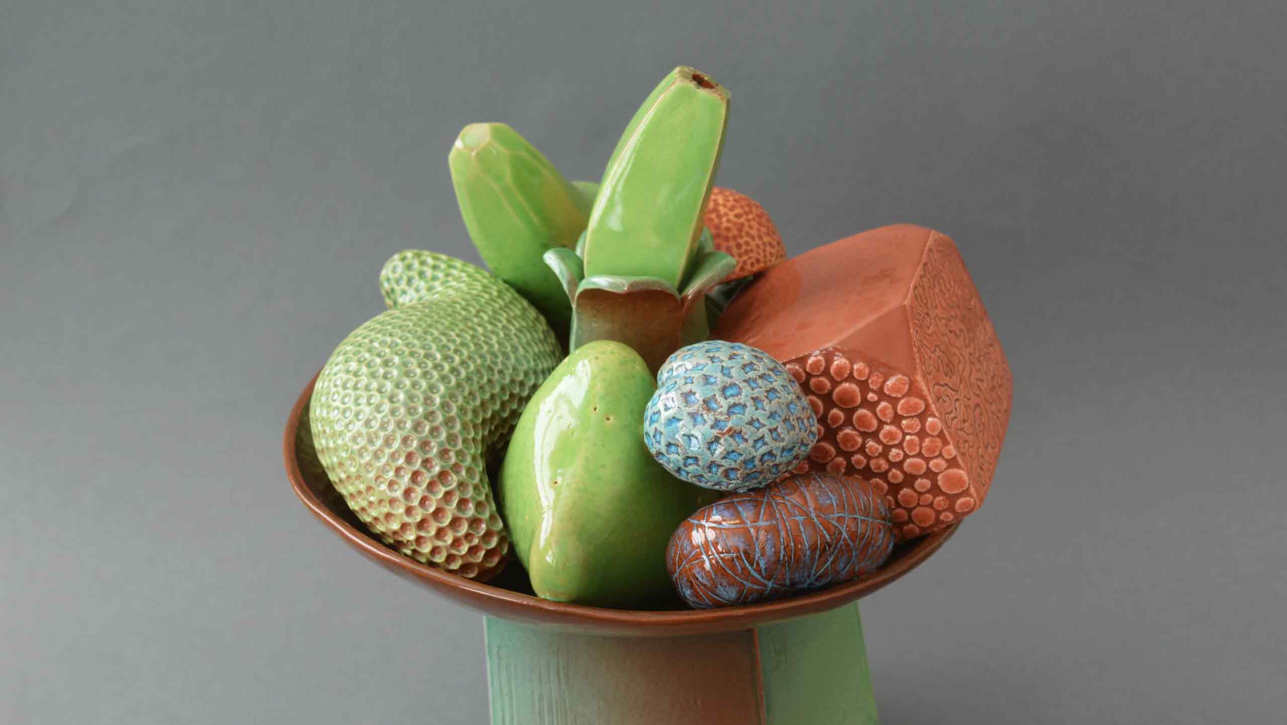 Photograph of 'Abundance' - still life on stand by contemporary ceramic artist Simon Fell: A ceramic still life on a bowl on a tall stand sits a set of unlikely fruits. Something like a banana emerges vertically from a green flower, a box of pointed succulents emerge from a circular opening, a green pear becomes crystalline at one endand a blue dimpled heart sits next to a double ended curvaceous avocado.
