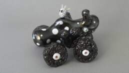 A four-wheeled figure in metallic black with white spots and sprigs of toothbrush hair. Ceramic earthenware 17cm long, 14cm wide, 14cm high
