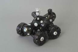 A four-wheeled figure in metallic black with white spots and sprigs of toothbrush hair. The wheels are made of compressed coils like the structure of a brain.