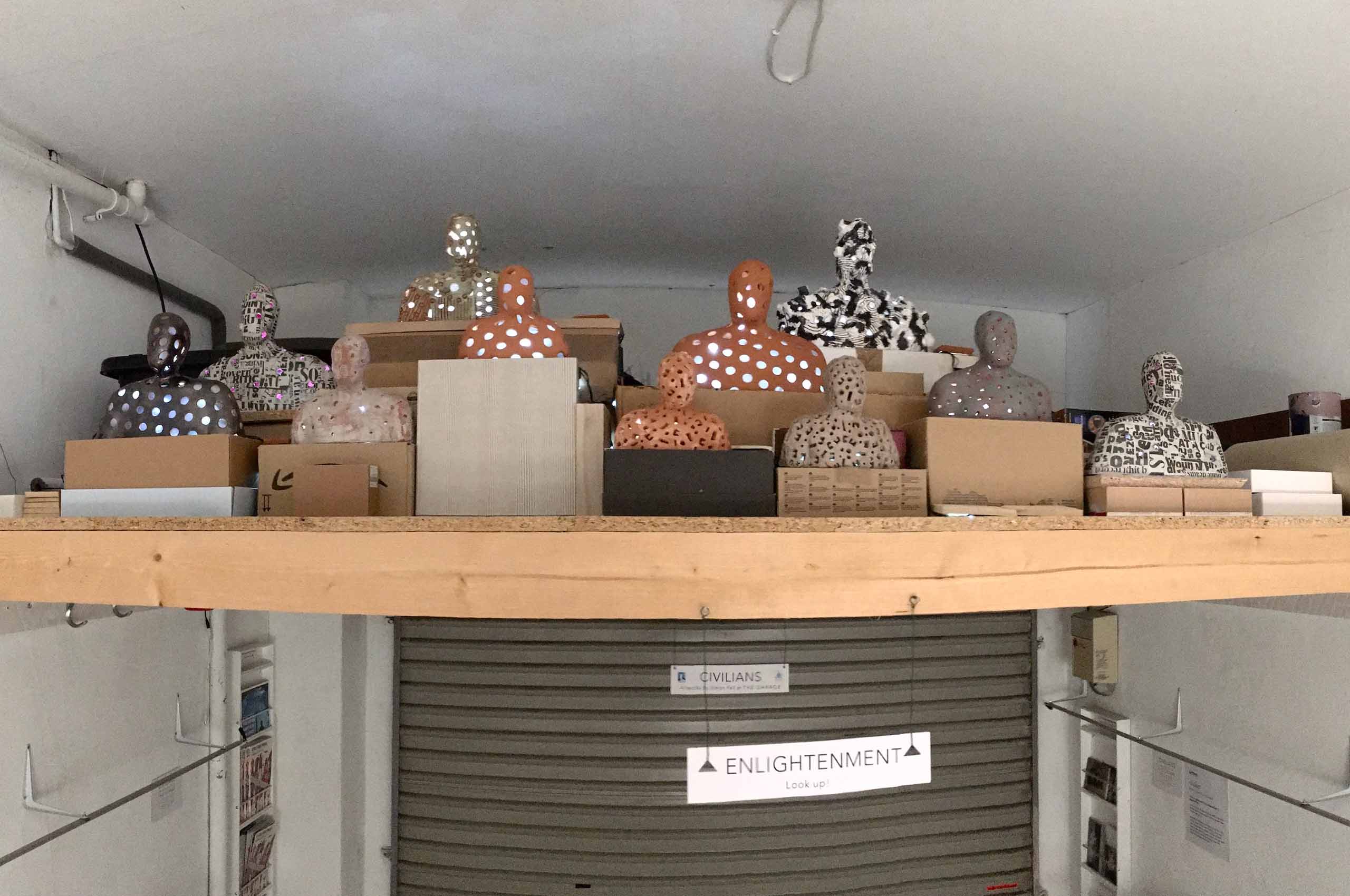 A daylight view of a developing installation of 13 or more figures in glazed and unglazed terra-cotta, plaster and papier mache illuminated by LED lights on stands made of cardboard and mdf