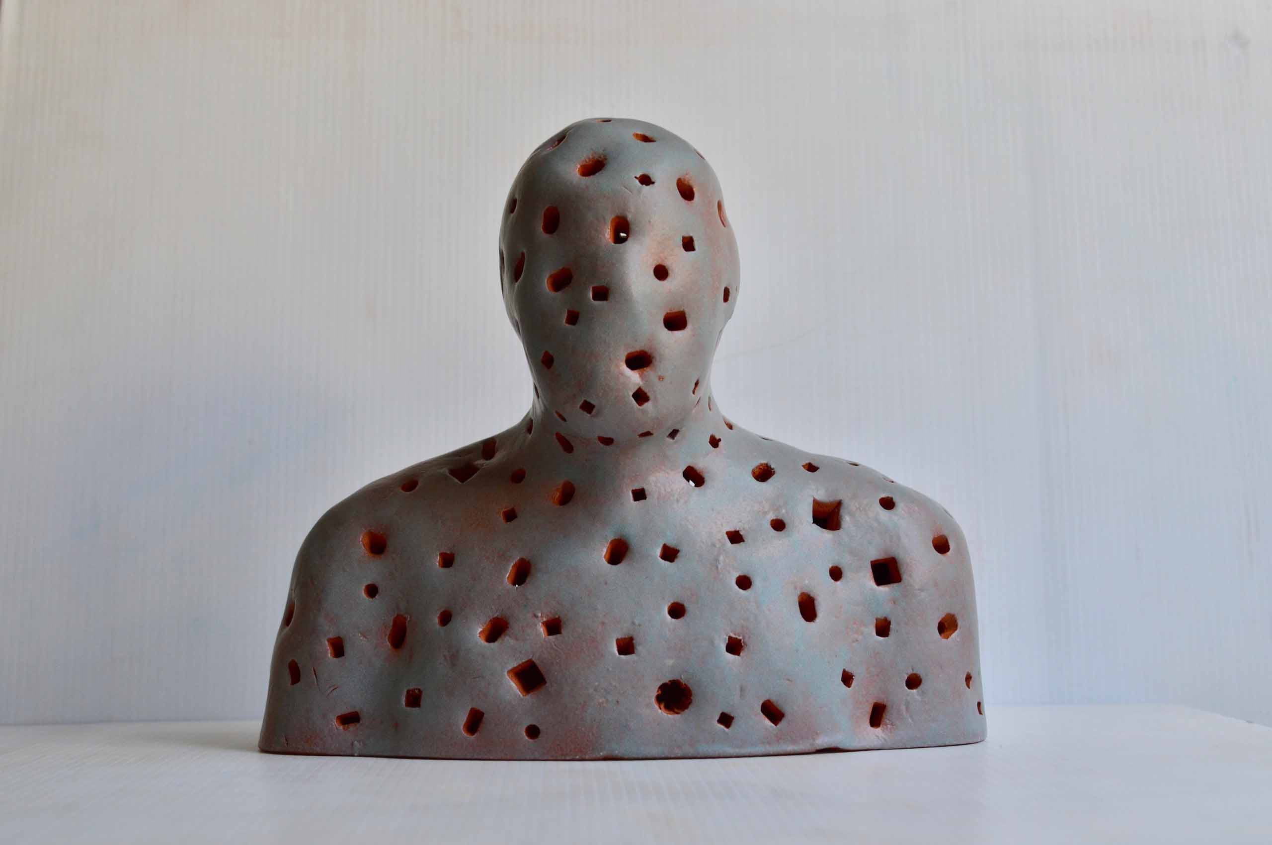 Ceramic sculpture of head and shoulders with stabbed holes of different sizes and shapes that reveal the thickness of the terra cotta clay it is made of. The figure is glazed in grey and turquoise