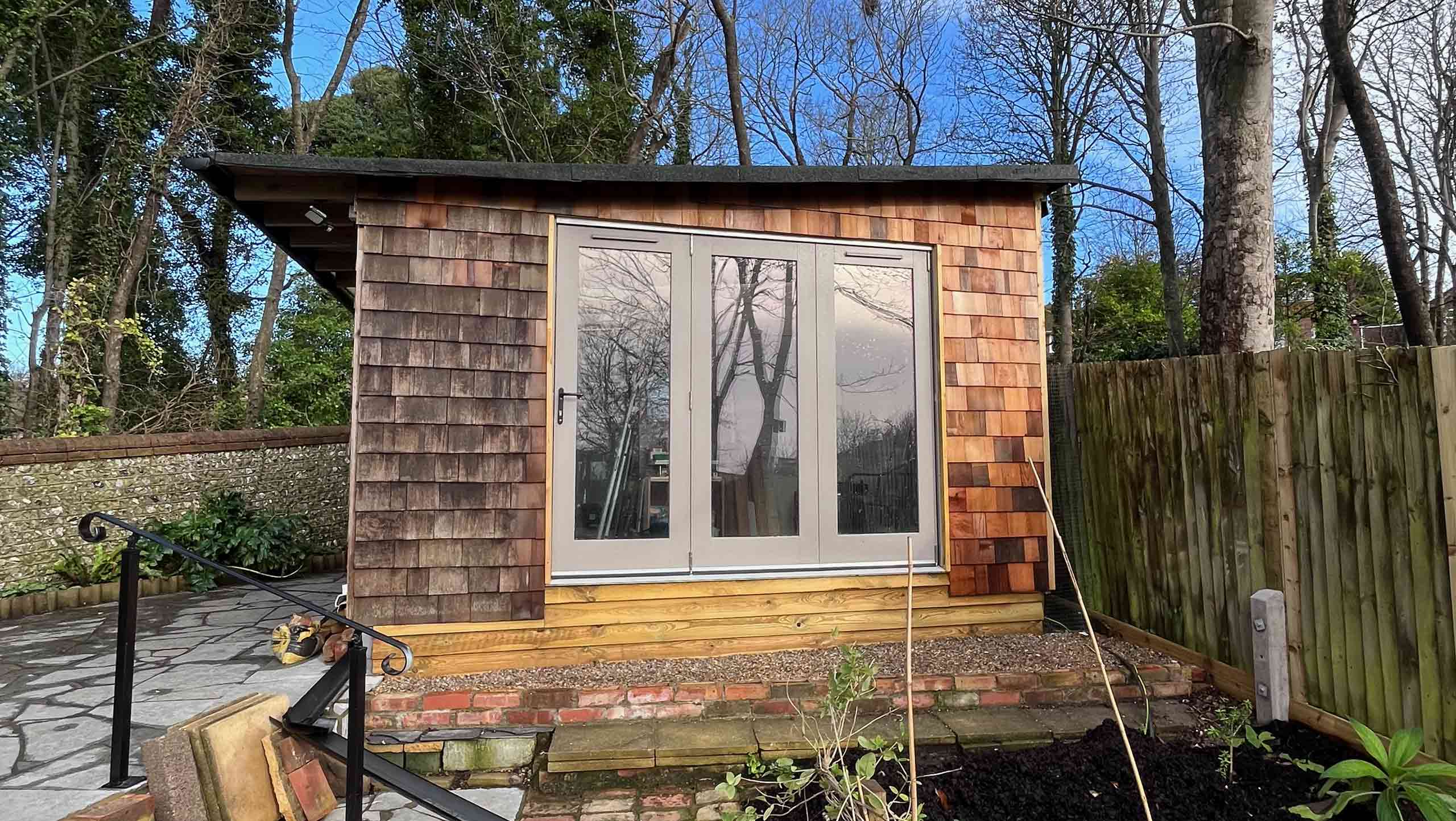 The studio after it has rained. The south end is largely made up of three french windows with grey frames. The rest of the end is shingled, these change colour in the rain becoming a richer tone of russet red.