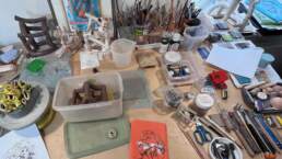 A wide desktop covered in tools, boxes, a turntable, several scupltures in different states and a lot of brushes and pencils in pots
