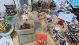 A wide desktop covered in tools, boxes, a turntable, several scupltures in different states and a lot of brushes and pencils in pots