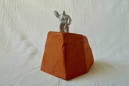 An aluminium figure, heavy set as if wearing a space suit or equally it could be bandaged, there are horizontal markings across his whole body from head to toe. The figure is cast in aluminium from a clay original and fettled by hand. It’s about 40mm high, the base is about 80mm high. An edition of five cast aluminium figures on a press moulded, terracotta base.