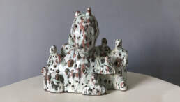 Ceramic sculpture of head and shoulders with smaller figures protruding. White glaze has been poured roughly over the whole group leaving patches of brown clay with tinges of copper green.