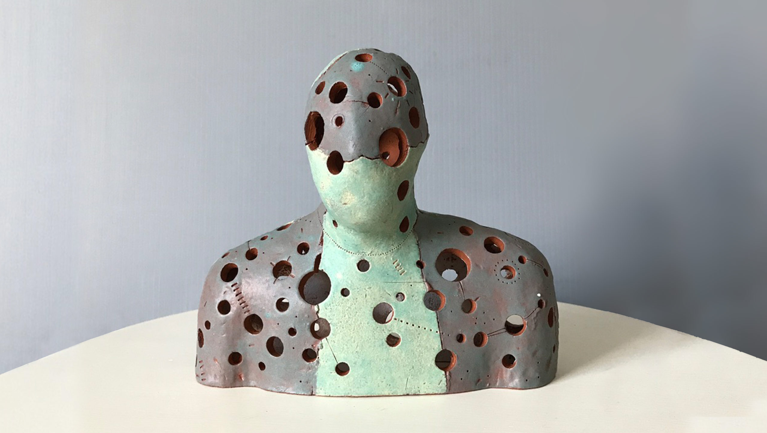 Ceramic sculpture of head and shoulders with circular holes of different sizes that reveal the thickness of the terra cotta clay it is made of. The figure is glazed in grey and turquoise in panels where it has been broken apart and re-assembled