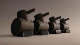 Tank Family - Four toy tanks lined up in a row in descending size order. Each tank is made of stoneware fired clay pinted with dark grey slip. They are propped up on grey glazed wedges and hold their guns up turned slightly away from the viewer as if they are saluting a passing general.