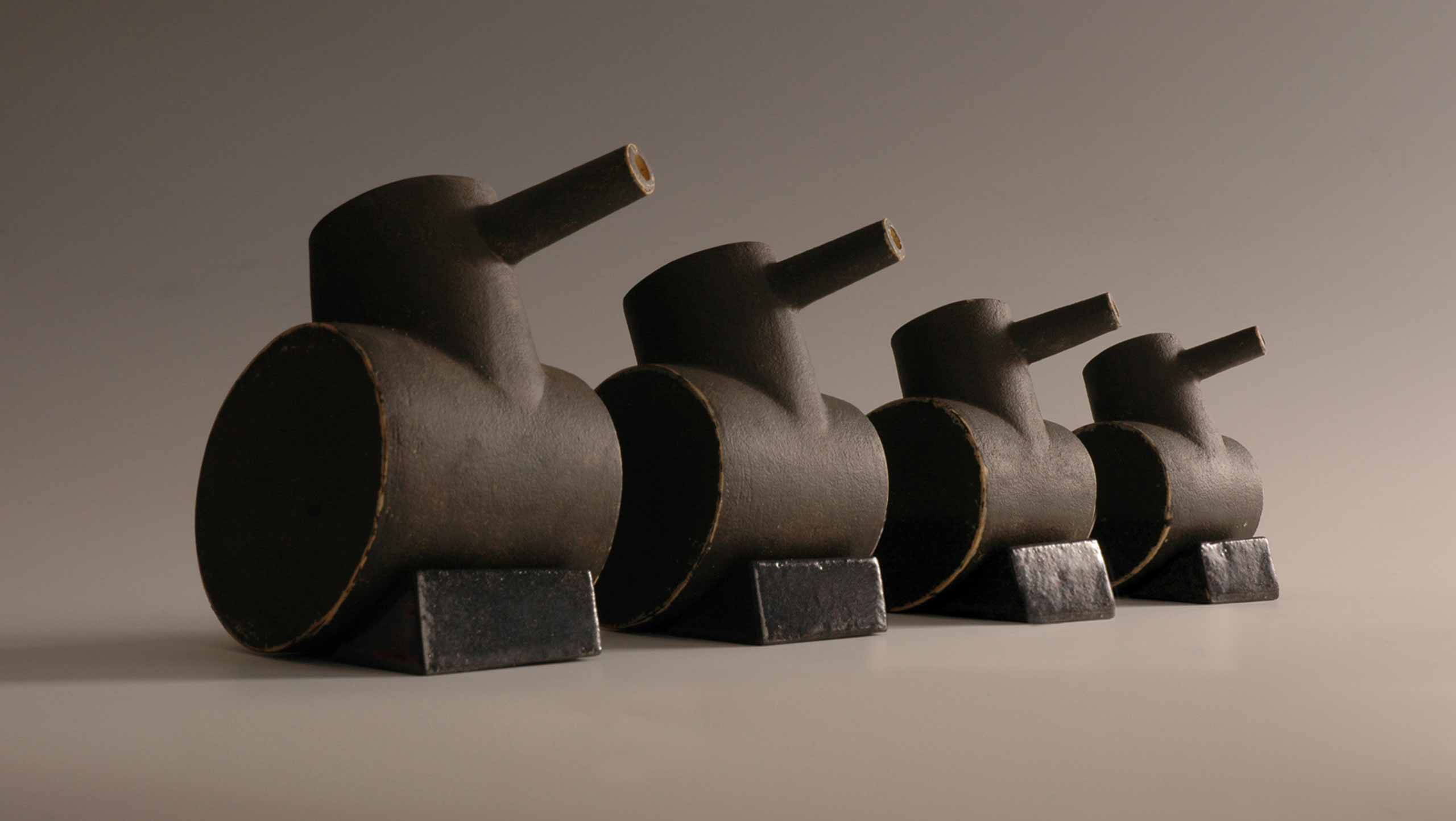 Tank Family - Four toy tanks lined up in a row in descending size order. Each tank is made of stoneware fired clay pinted with dark grey slip. They are propped up on grey glazed wedges and hold their guns up turned slightly away from the viewer as if they are saluting a passing general.