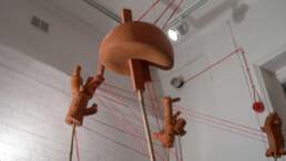 Tension-extension a ceramic installation by Simon Fell at Redlees Studios. Articulated terra-cotta forms are connected with red thread. Each form stand on a bamboo pole stuck into a black cupboard