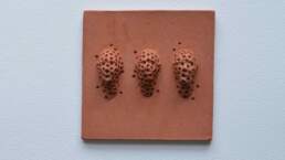 Tile with 3 heads, 10cm square terra-cotta tile unglazed. Showing 3 faces protruding textured or perhaps defaced with cross shaped indents