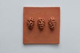 Tile with 3 heads, 10cm square terra-cotta tile unglazed showing 3 faces protruding textured or even defaced with square indents