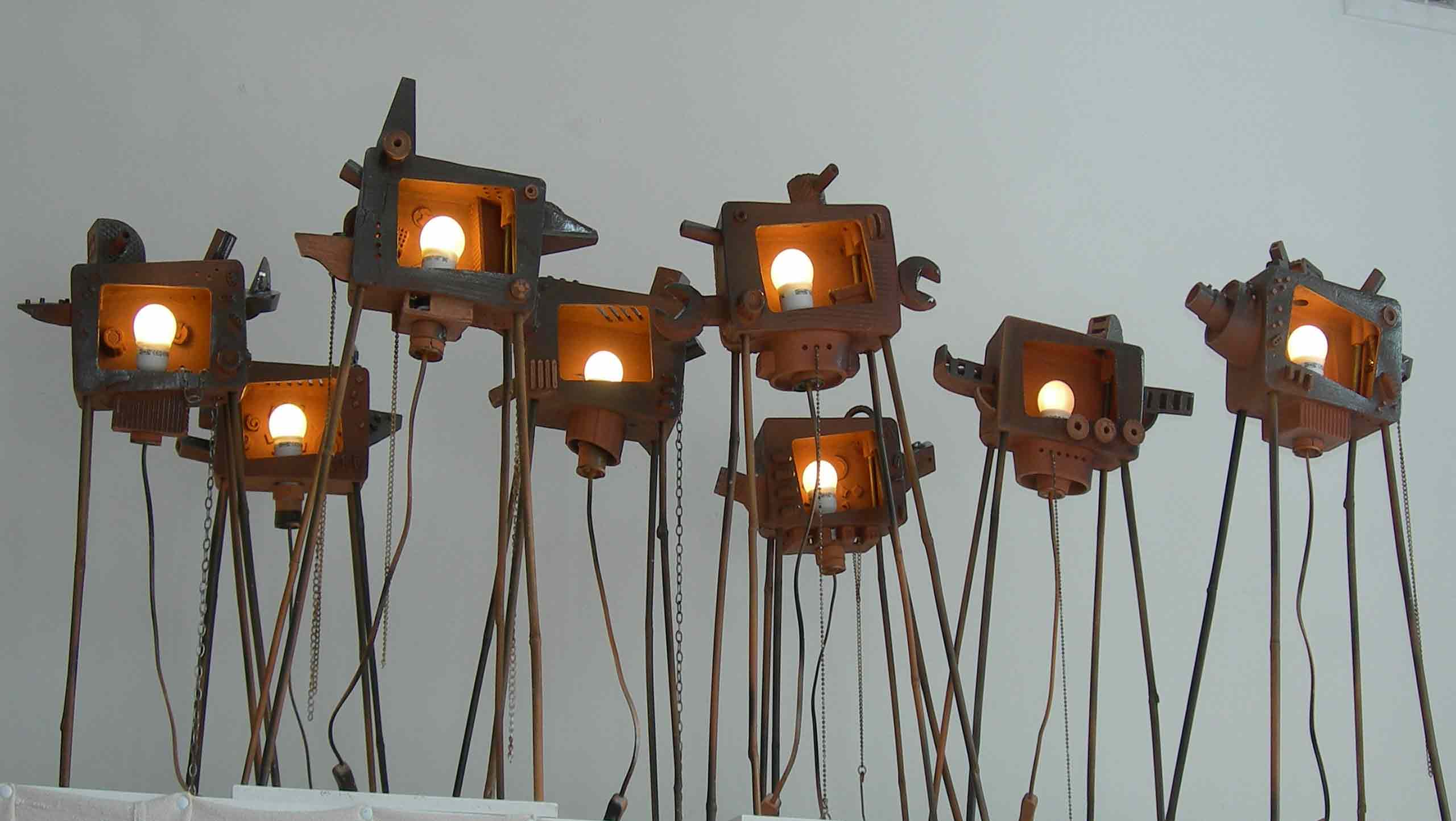 War of the Worlds, a ceramic installation by Simon Fell contemporary ceramics: A group of eight TV sets on tall thin stick legs with wires dangling stand in a line as if watching or guarding the way. Each set has attachments of unknown purpose that extend out of the basic TV shape. Each TV contains a bare frosted illuminated light bulb.